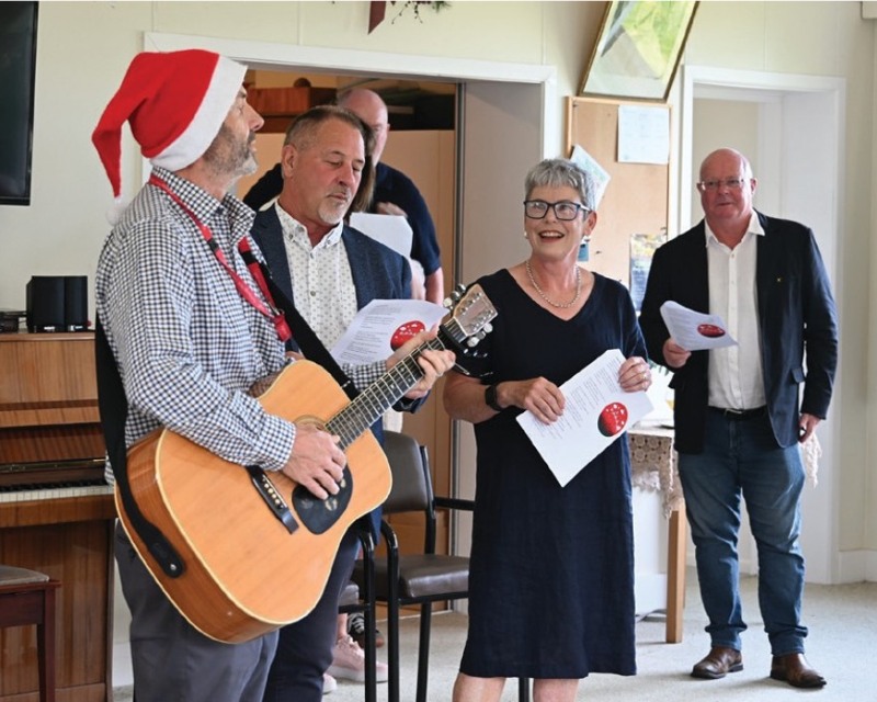 Four people including the Mayor sing Christmas carols as one person wearing a santa hat plays guitar